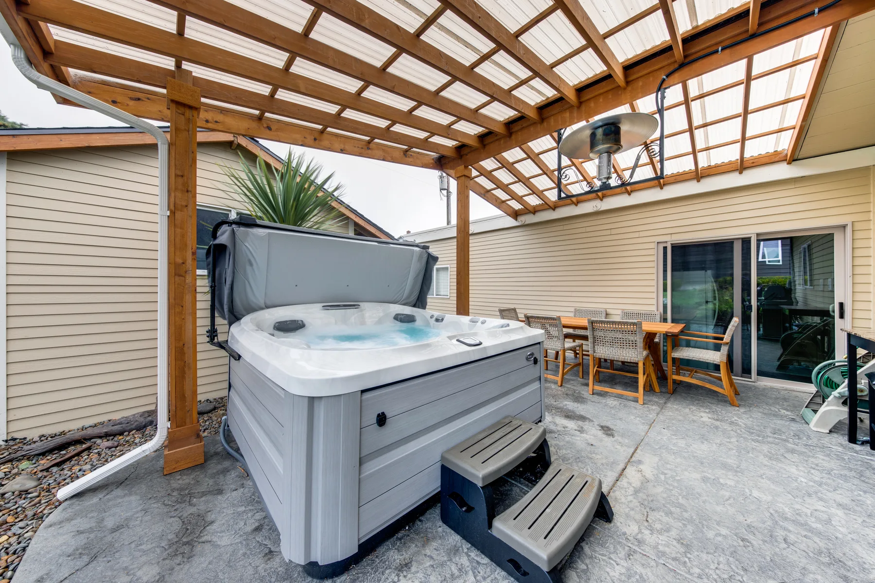 An outdoor hot tub and dining area sit beneath a covered wooden patio.