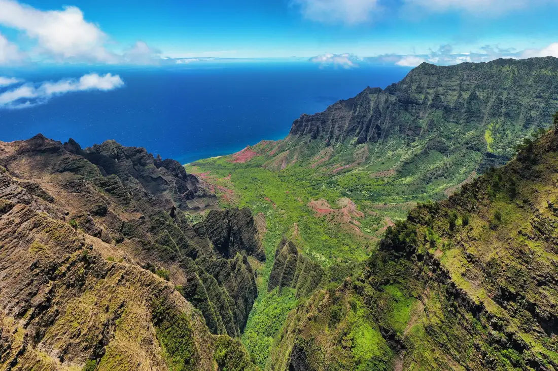 A green mountain valley opens toward the blue ocean beneath a bright sky.
