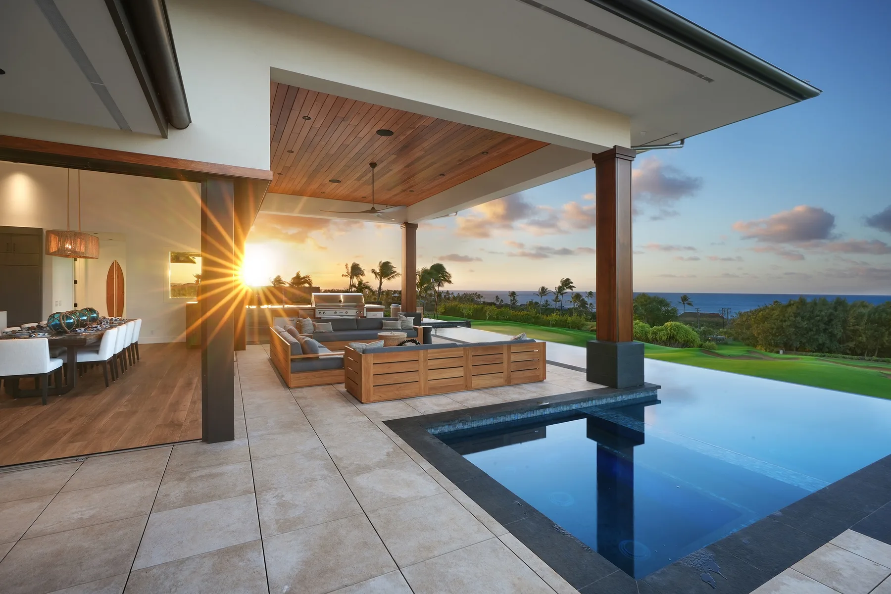 Covered patio with outdoor seating beside a pool overlooking the ocean at sunset.