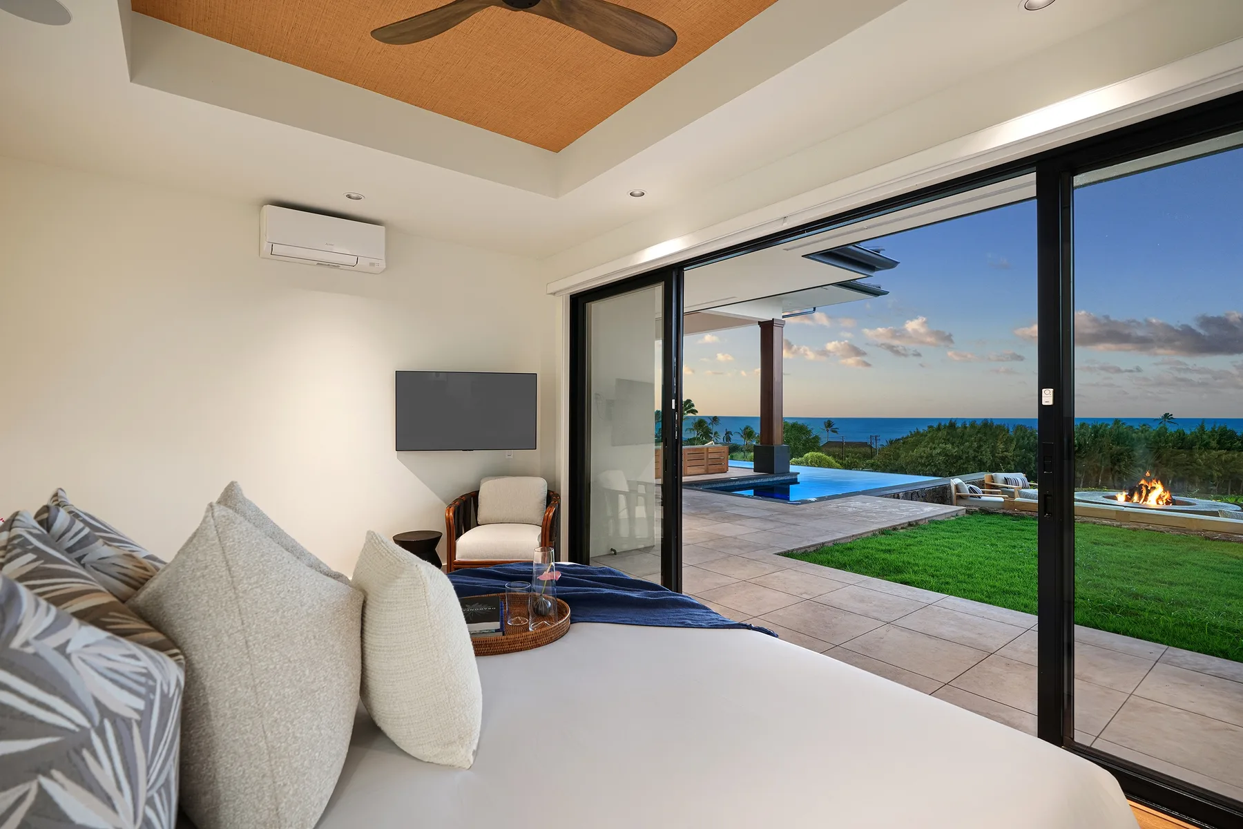 A bedroom opens through glass doors to a patio, lawn, and ocean view.