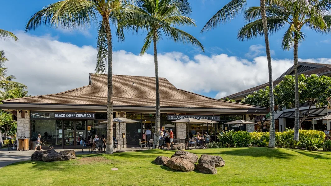A low-rise shopping complex with storefronts sits behind a lawn and palm trees.