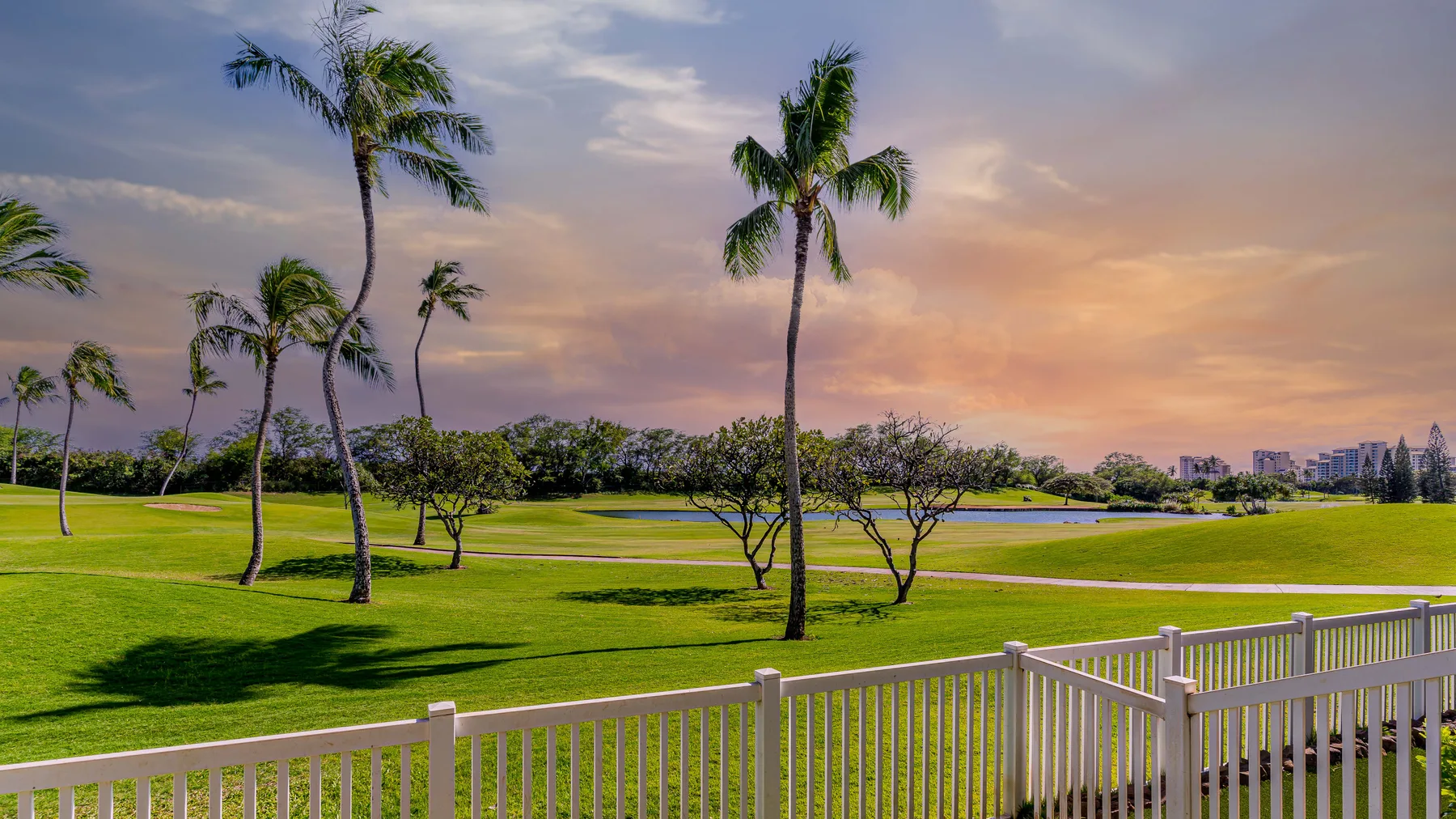 A landscaped golf course with palm trees and a white fence beneath a colorful sky.