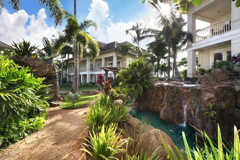 Un patio ajardinado con un camino sinuoso, plantas tropicales y un estanque junto a edificios de varias plantas.