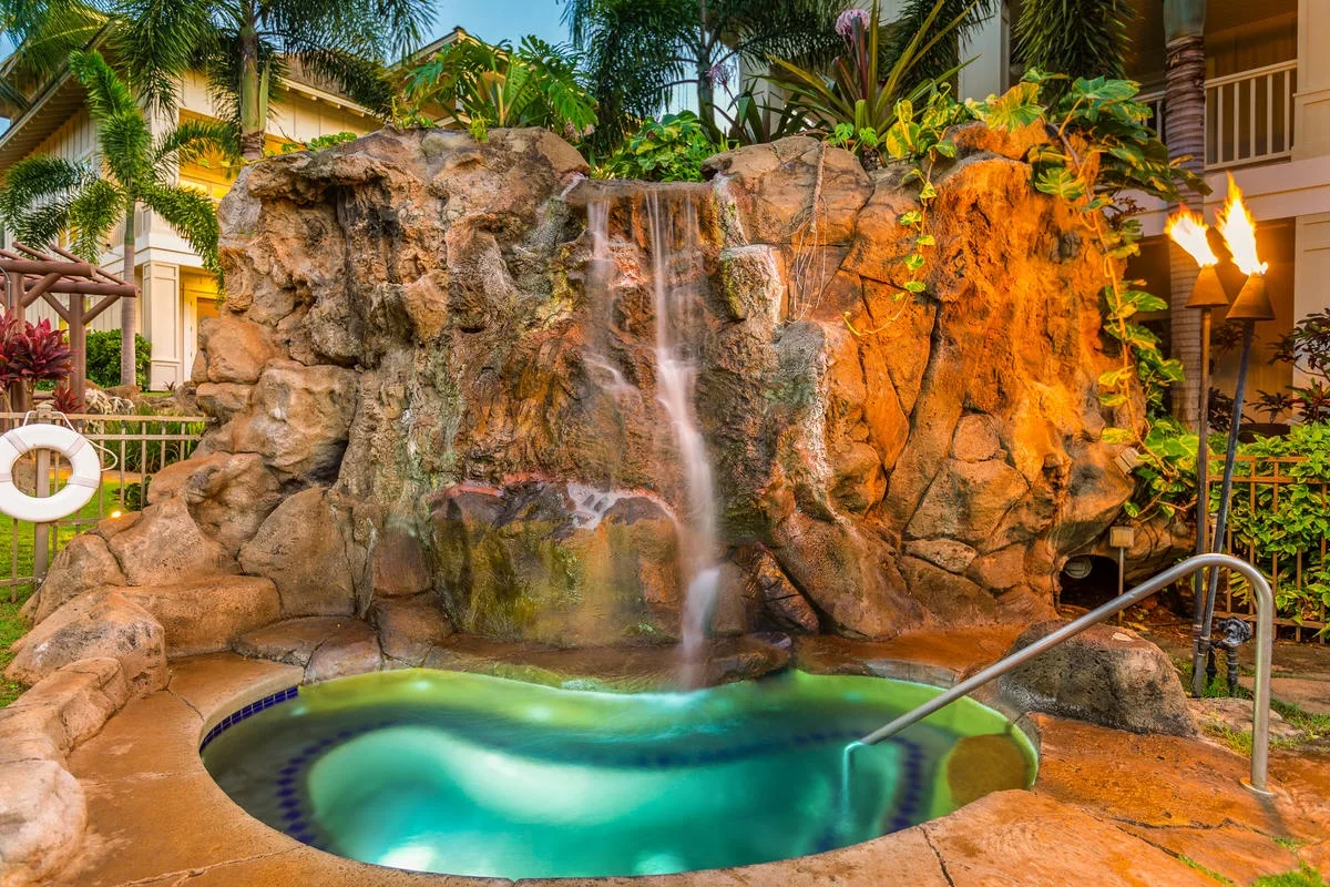 Una pequeña piscina está bajo una cascada rocosa en un jardín tropical.