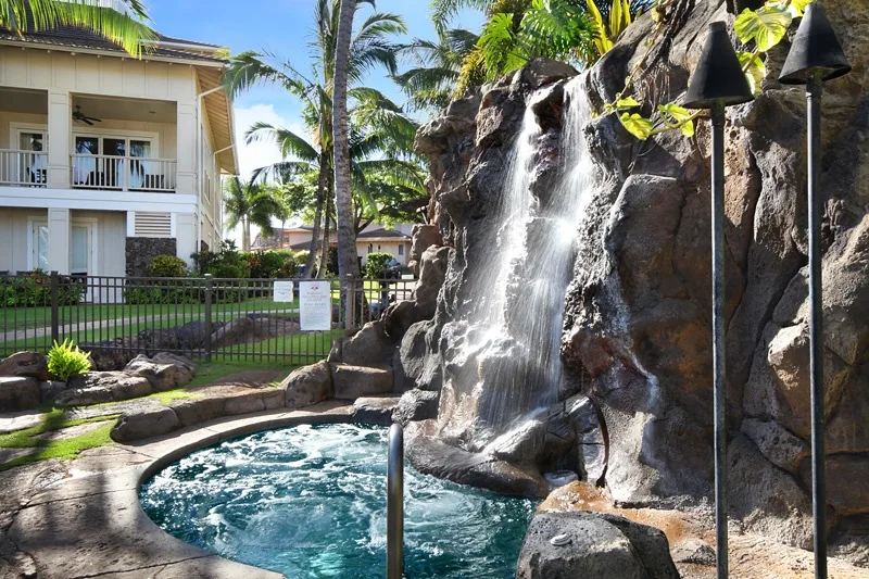 Una piscina exterior burbujeante está junto a una cascada rocosa, con palmeras cerca.