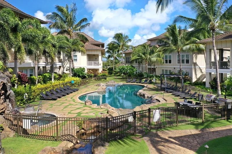 Una piscina tropical está rodeada de palmeras, asientos para descansar y edificios.