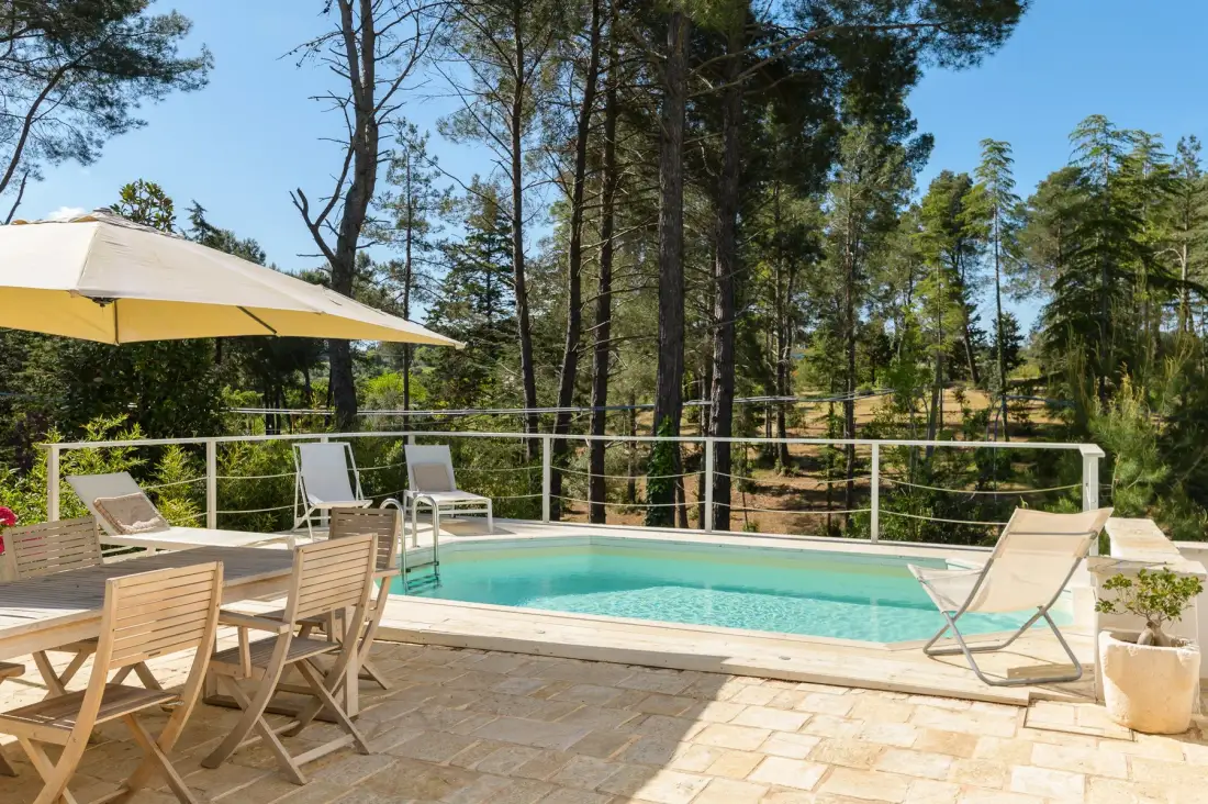 Una piscina al aire libre está rodeada por un patio pavimentado y árboles.