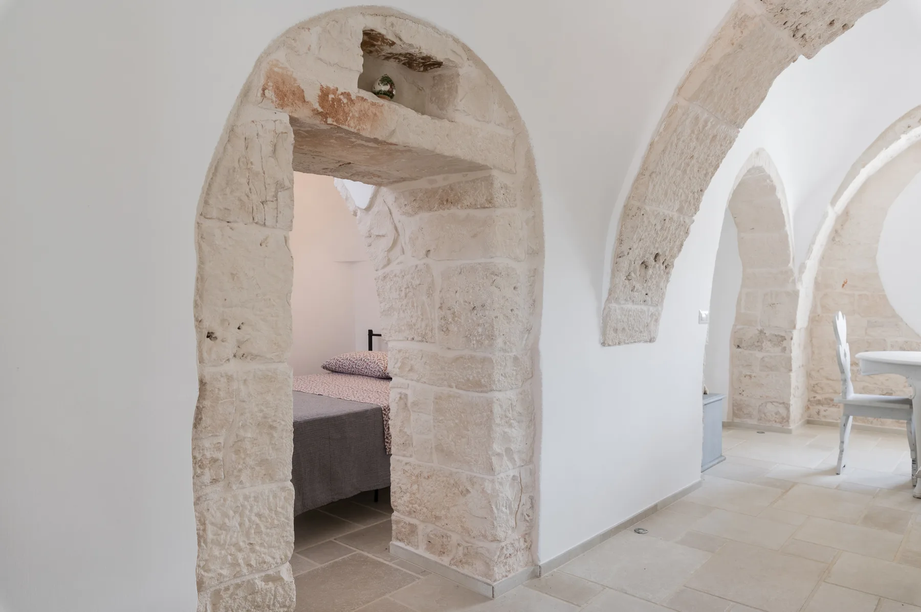 Se ve un dormitorio a través de un arco de piedra junto a un pasillo luminoso con arcos.