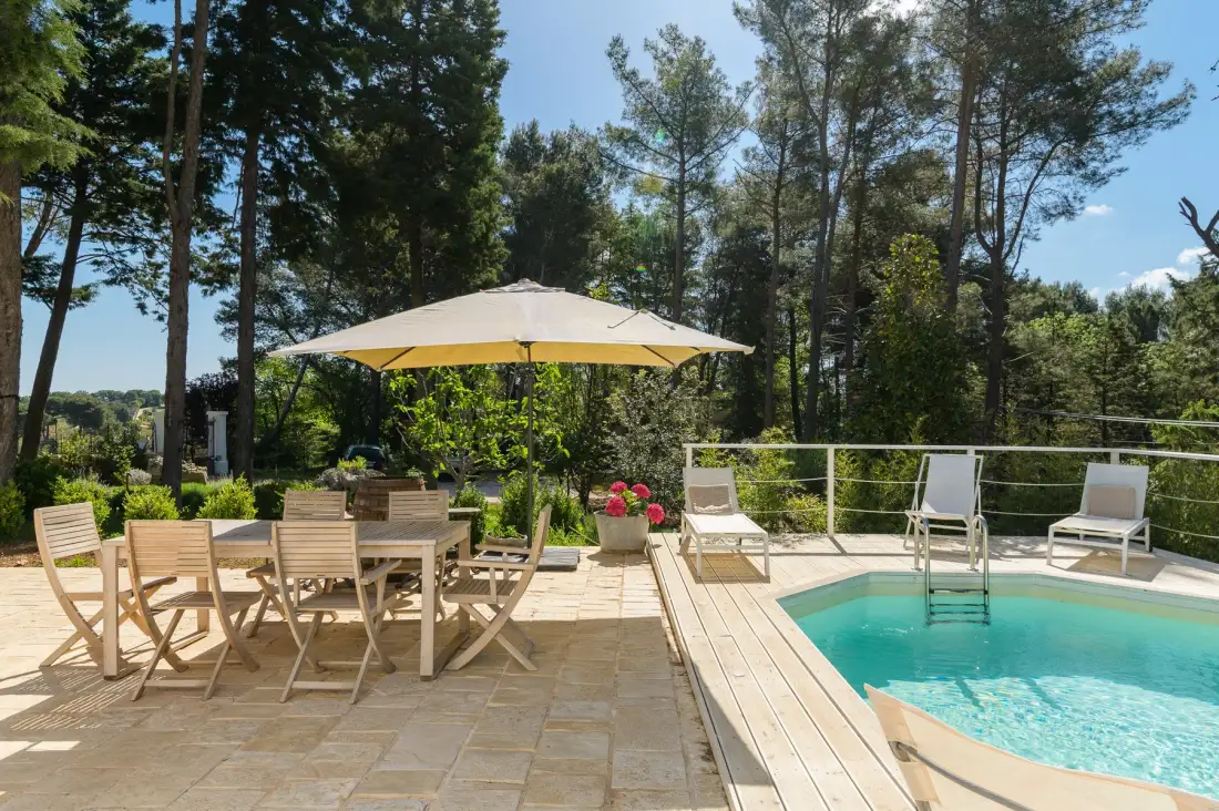 Una piscina exterior y asientos de patio están bajo una sombrilla grande, entre árboles.
