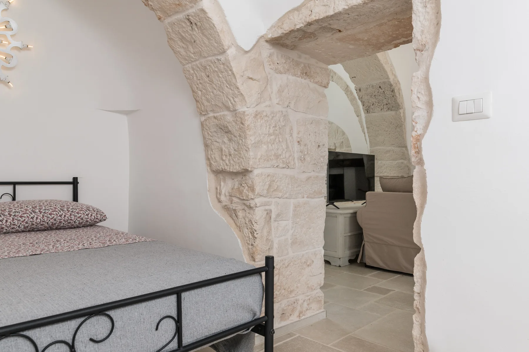 Un dormitorio con una cama metálica junto a un arco de piedra que da a una zona de estar.