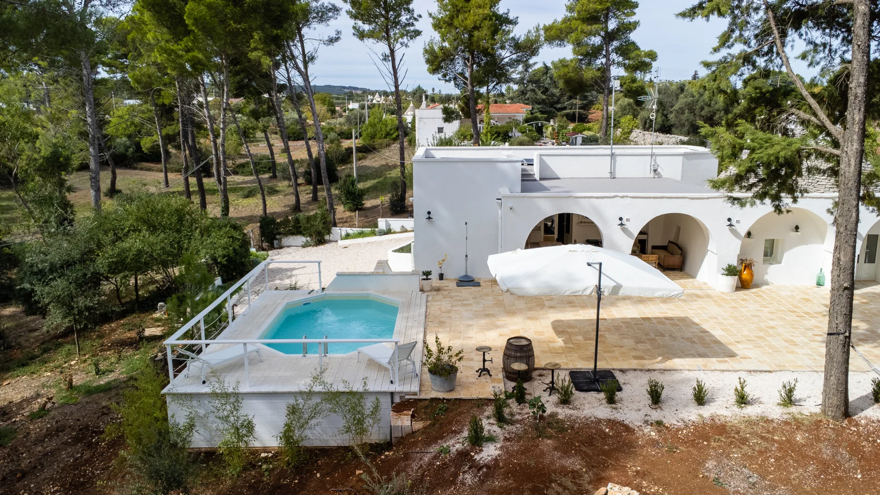 Una villa blanca con aberturas arqueadas da a una pequeña piscina y una terraza de piedra entre árboles.
