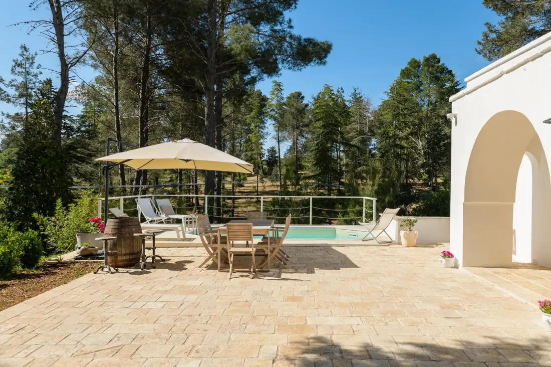 Una soleada terraza junto a la piscina, con asientos, una sombrilla y árboles al fondo.