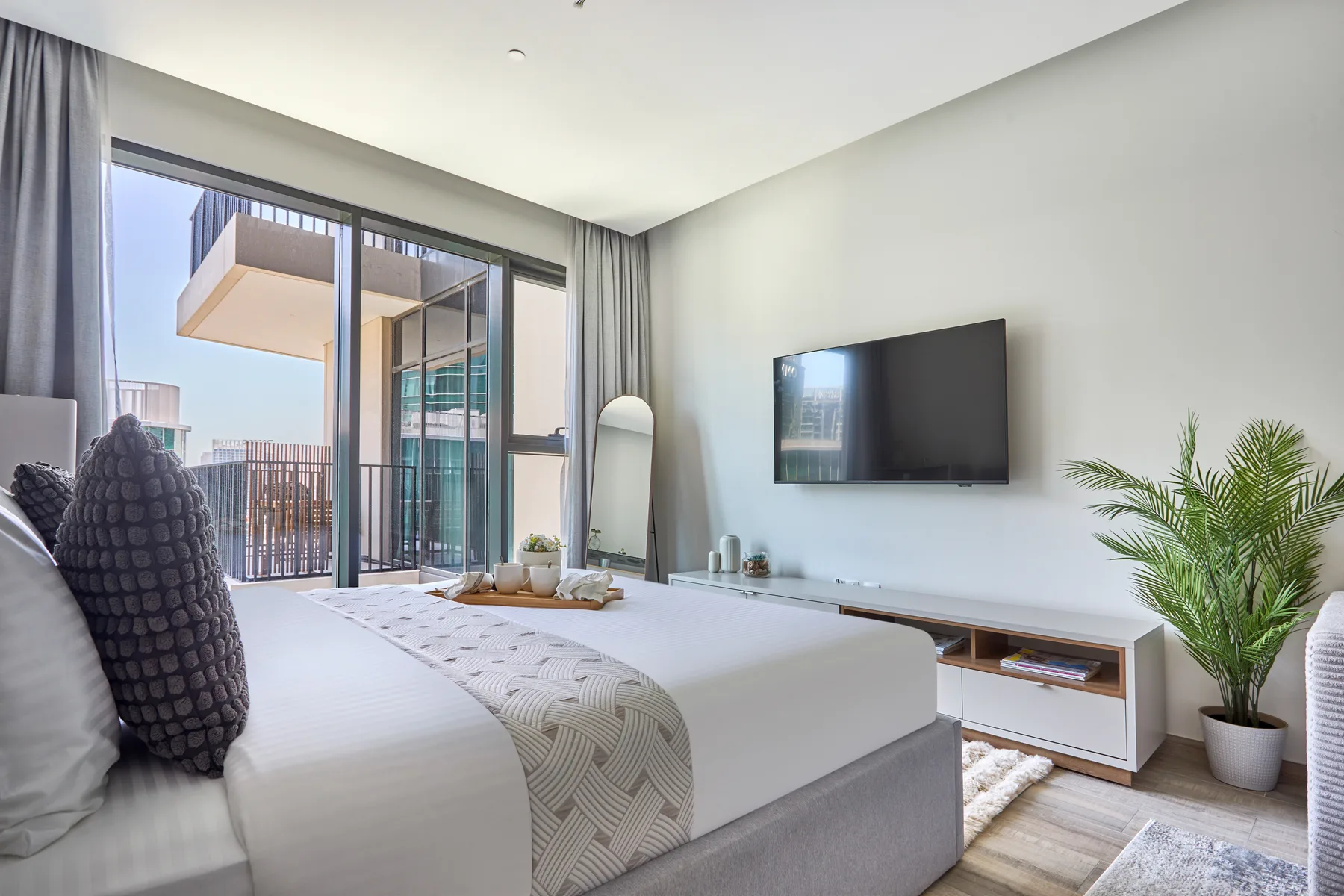 Un dormitorio luminoso con una cama arreglada, un televisor en la pared y una planta en maceta.