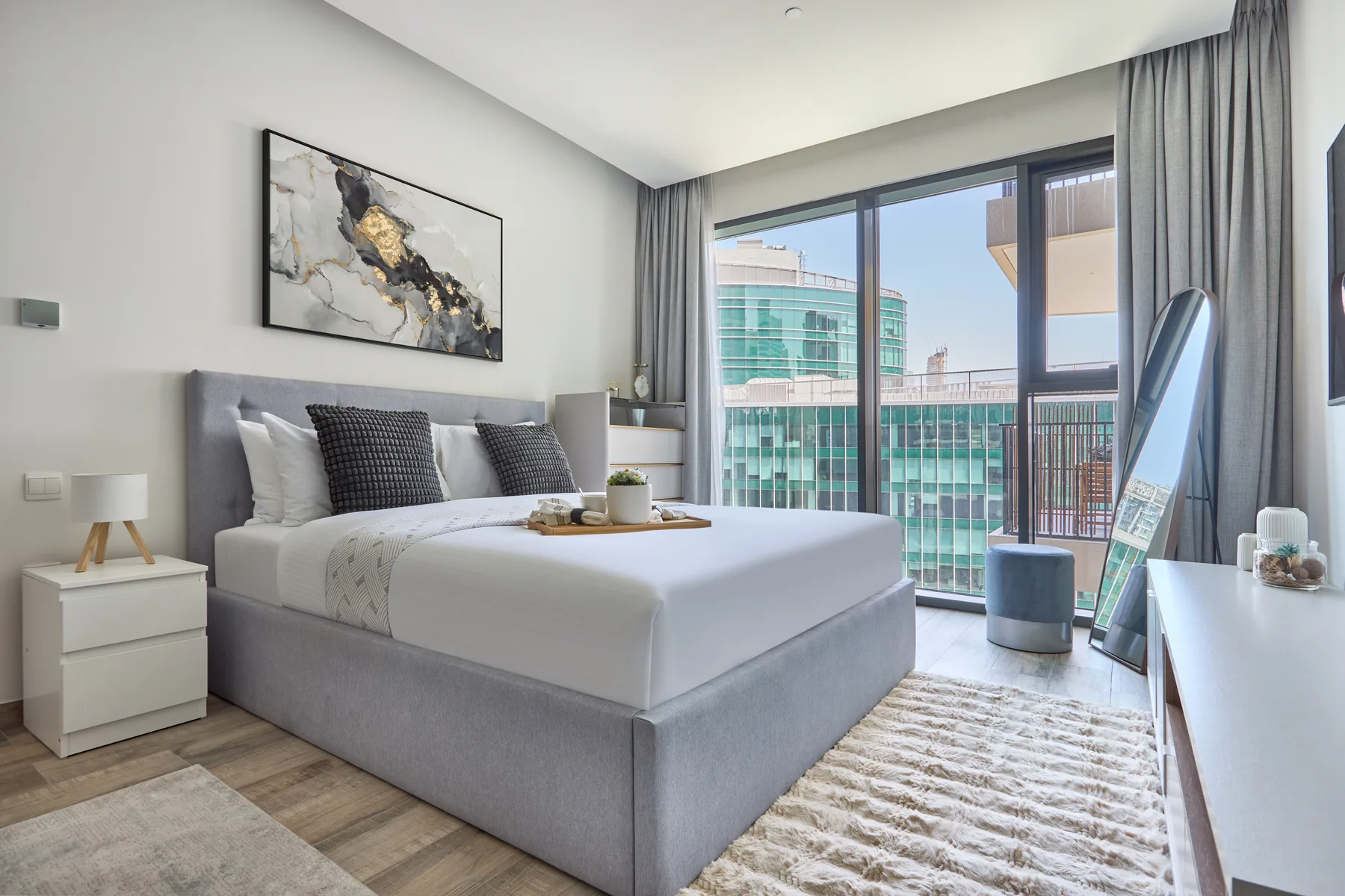 Un dormitorio luminoso con una cama bien hecha junto a grandes ventanas y cortinas grises.