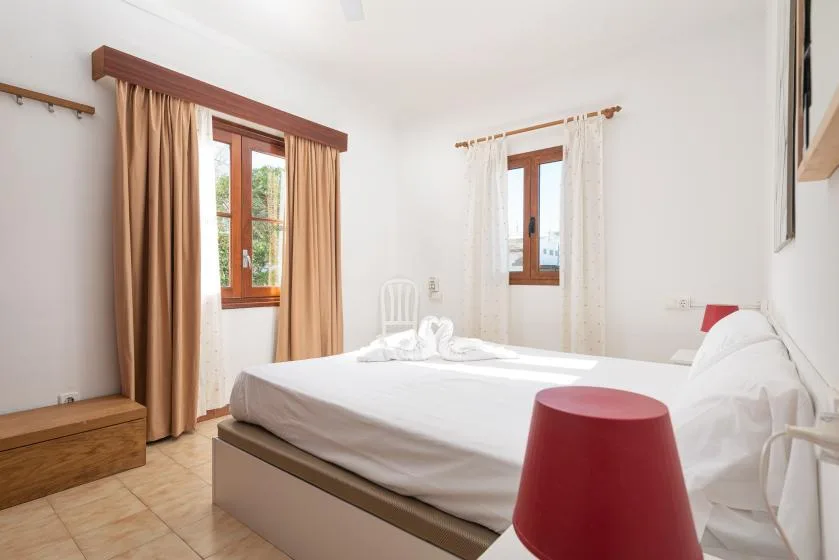 A bright bedroom with a bed, curtained windows, and a red bedside lamp.