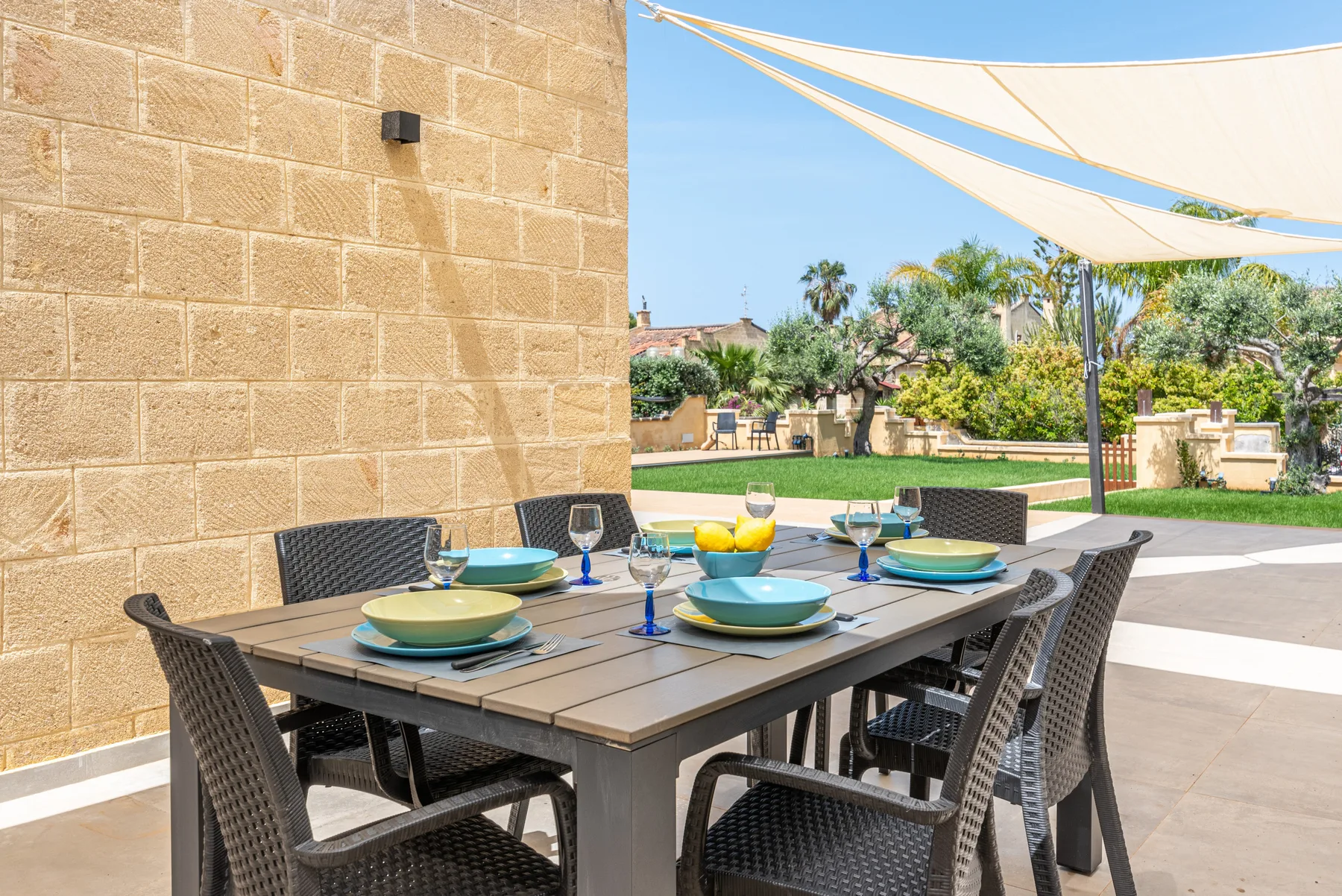Una mesa de comedor al aire libre está puesta con platos bajo una lona de sombra.
