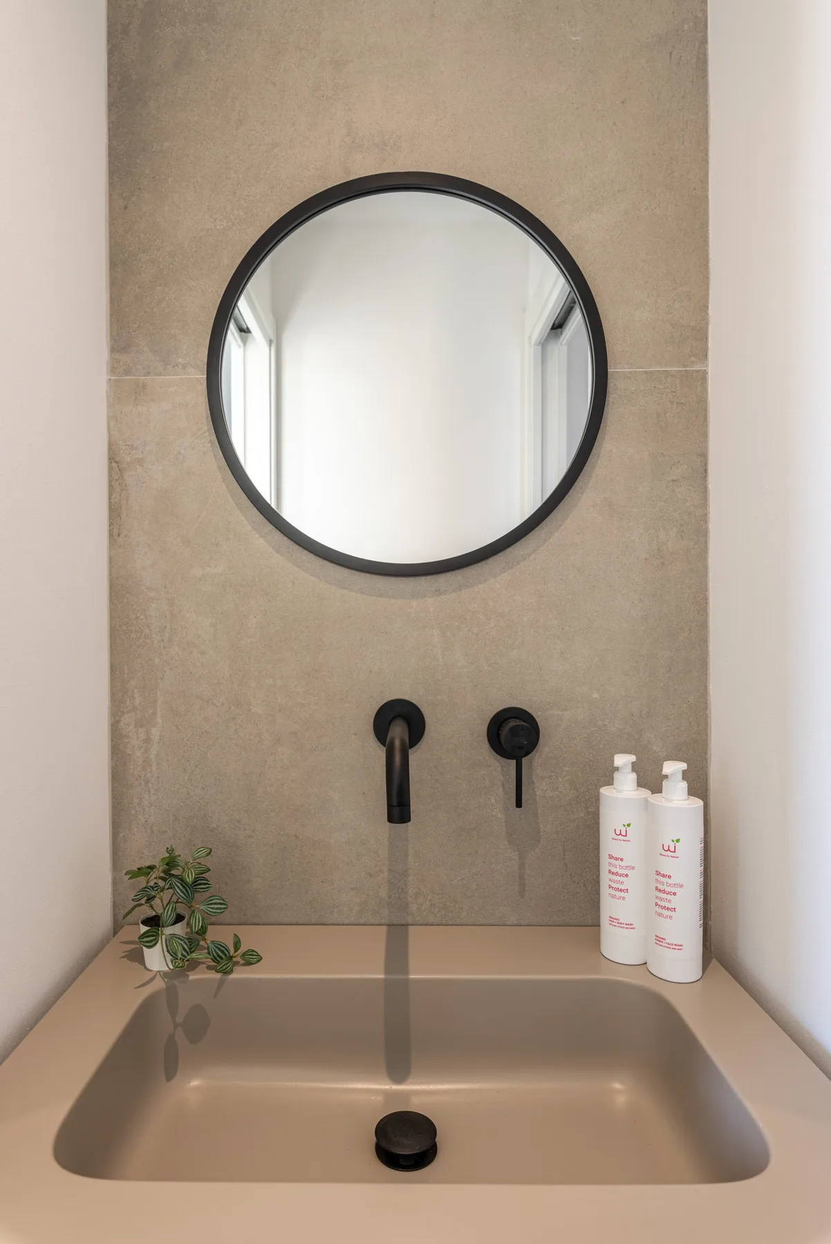 Un lavabo de baño con espejo redondo, grifería oscura y productos de aseo junto al lavamanos.
