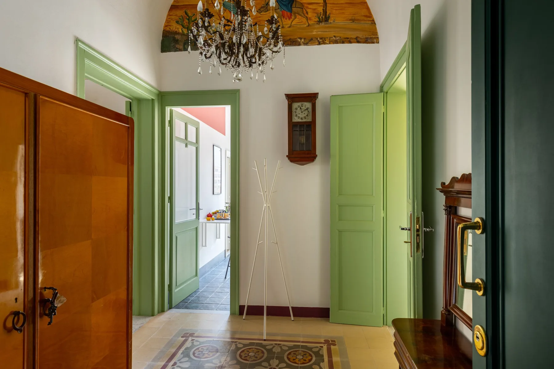 Un recibidor luminoso con puertas verdes, una lámpara de araña y una alfombra estampada.