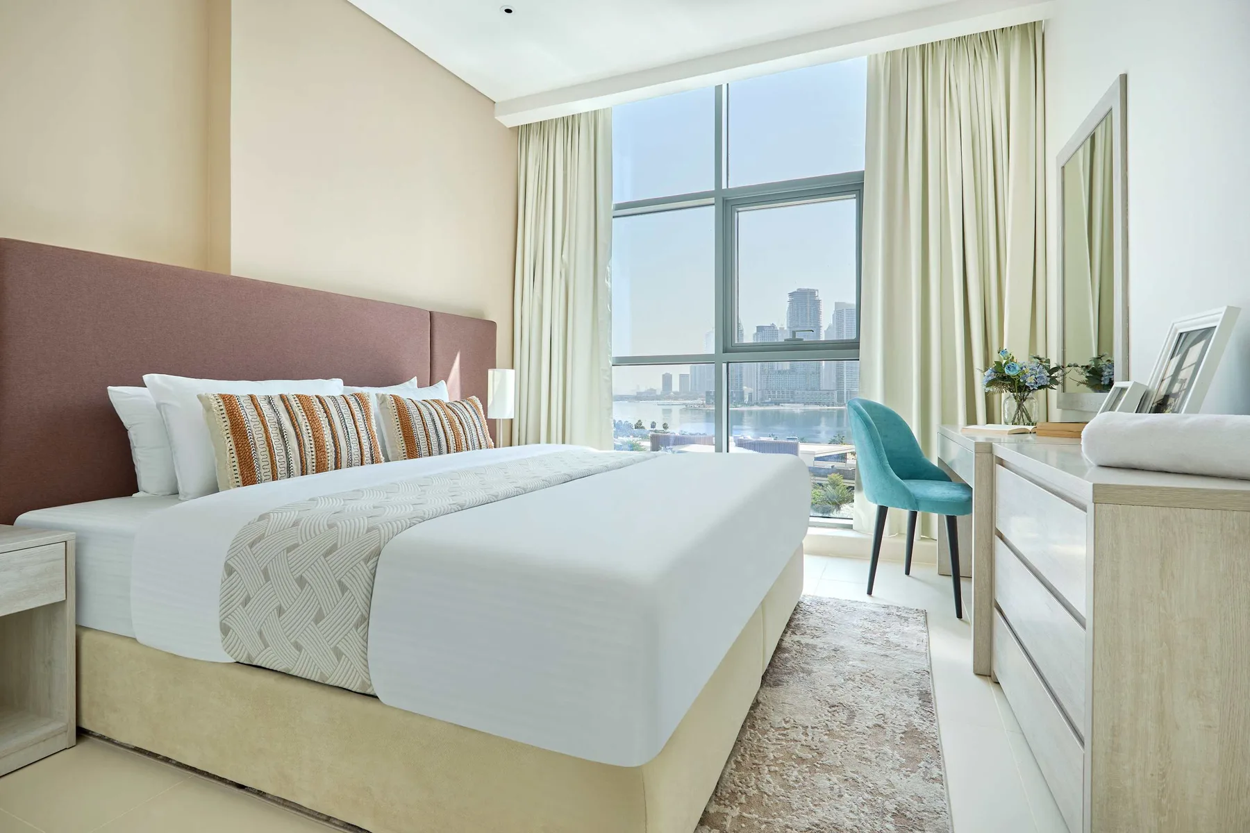 Un dormitorio luminoso con una cama grande, asiento turquesa y una ventana de piso a techo.