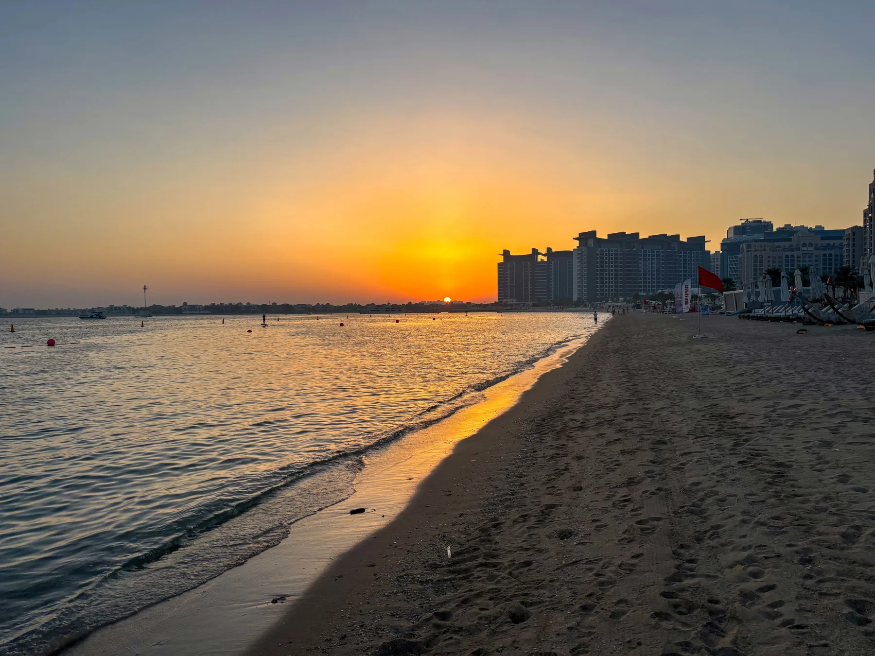 Una playa de arena al atardecer, con olas suaves y edificios junto a la costa.