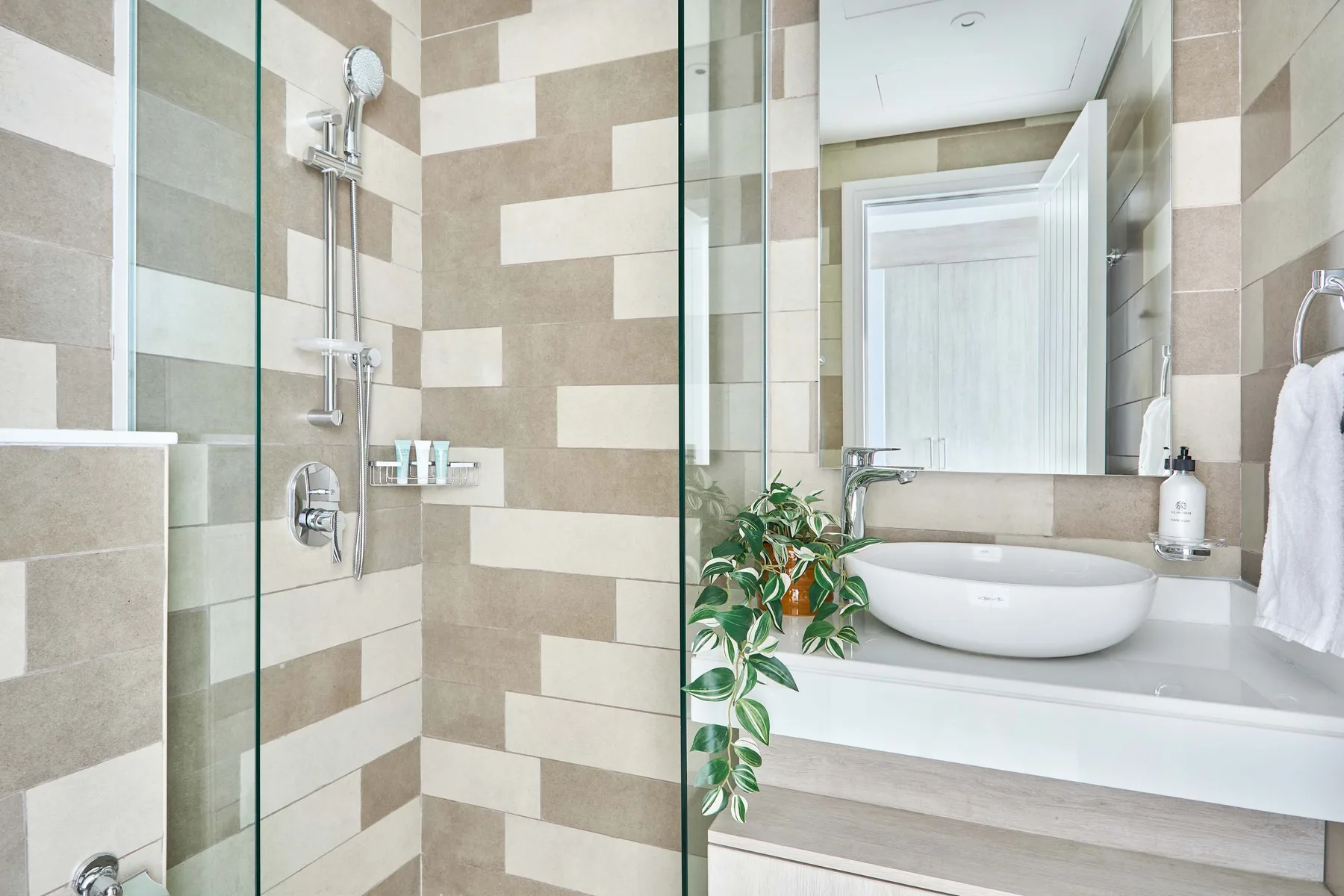 Baño alicatado con ducha de vidrio, lavabo blanco y una planta frondosa.