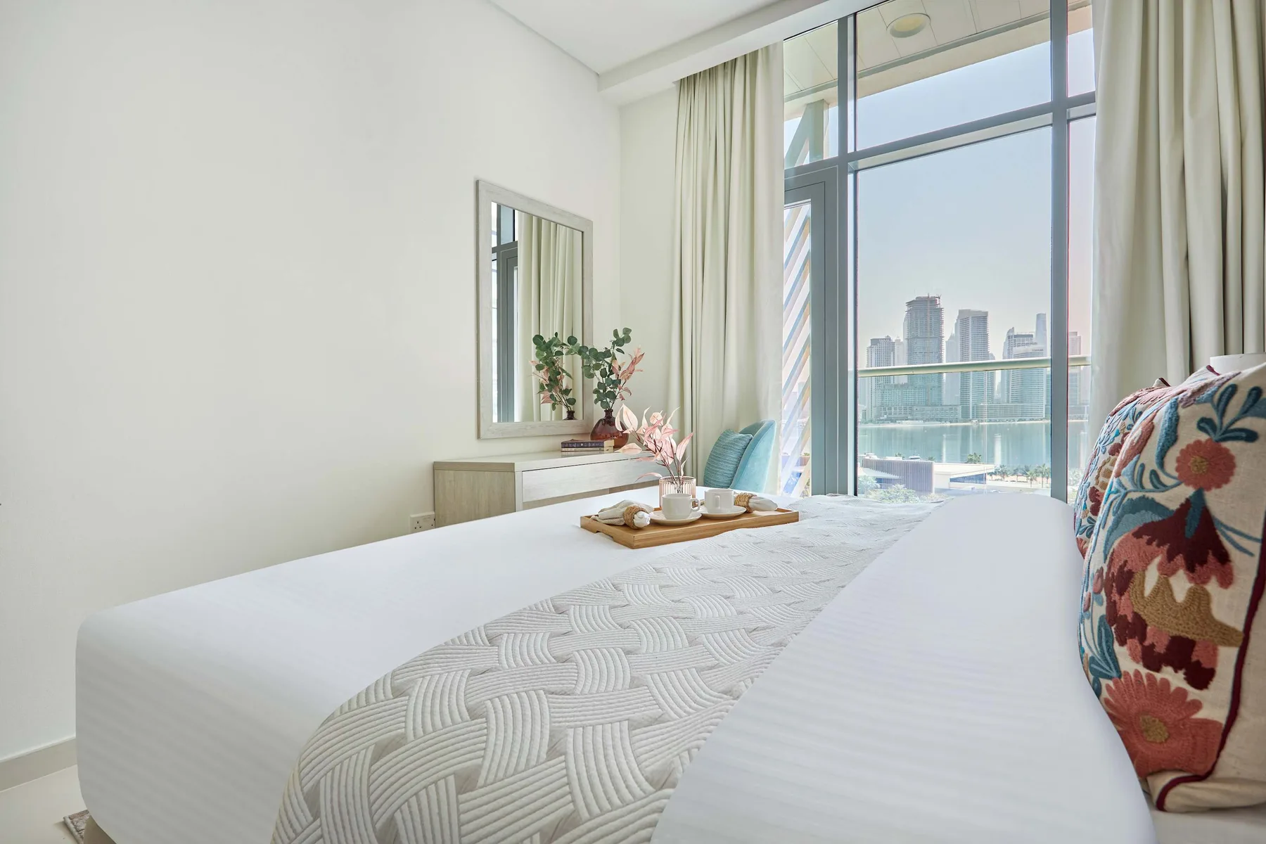 Un dormitorio luminoso con una cama arreglada junto a grandes ventanas y cortinas claras.