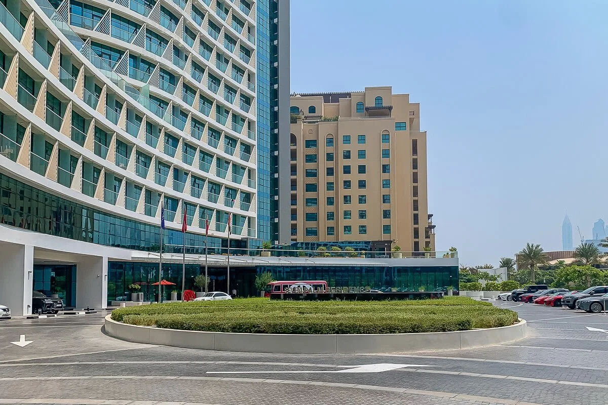 Edificios modernos de gran altura dan a una entrada circular ajardinada con autos estacionados.