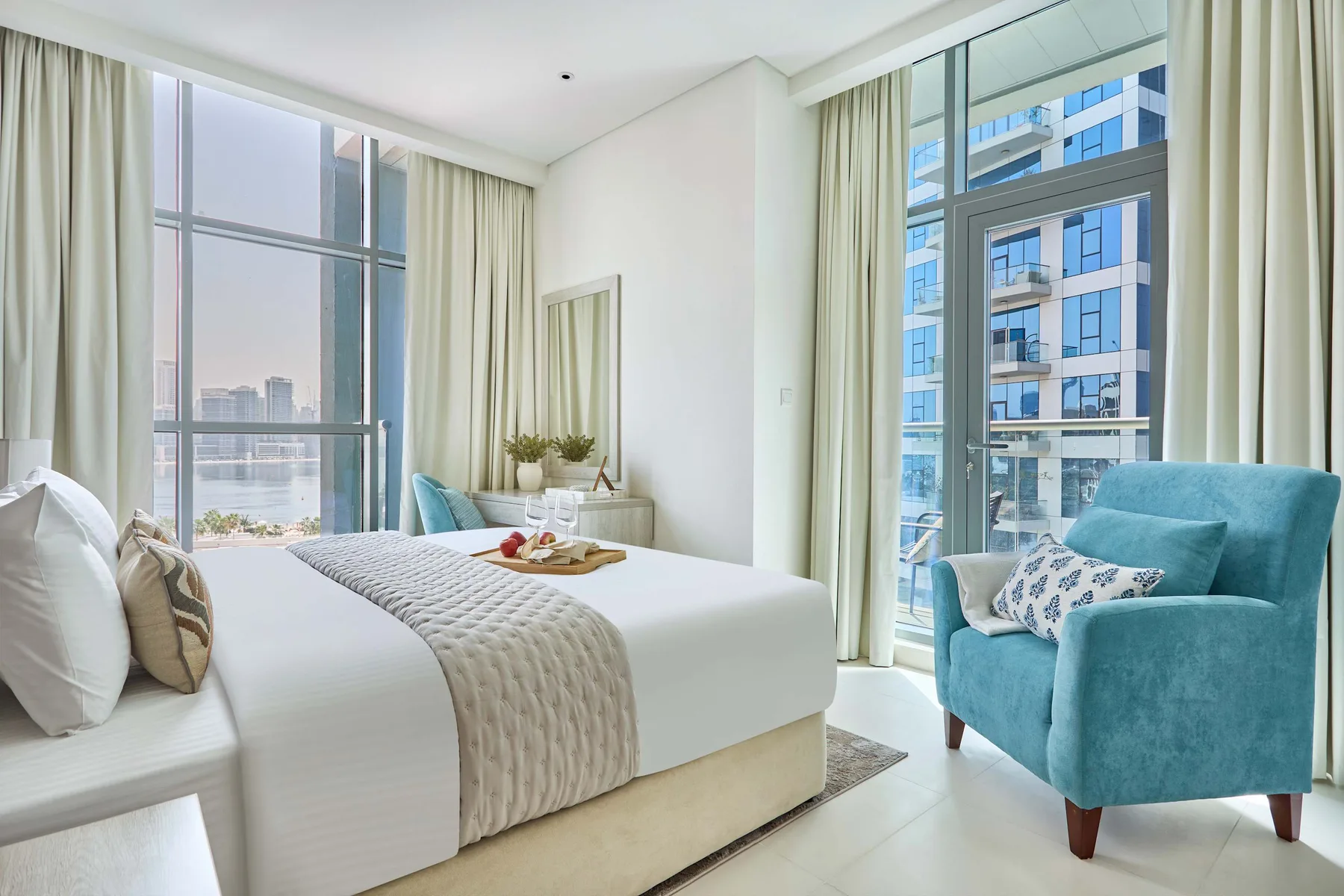 Un dormitorio luminoso con una cama bien hecha y un sillón turquesa junto a grandes ventanas.