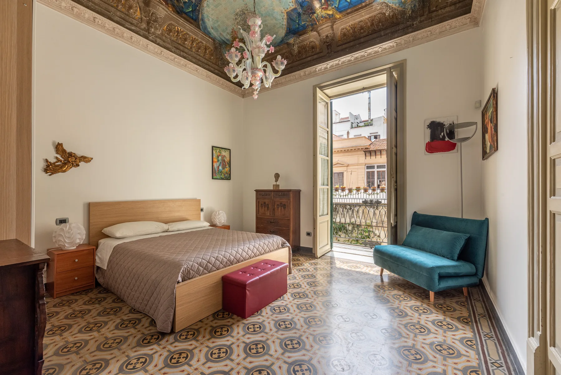 A bedroom with a bed, a teal armchair, and an open balcony door.