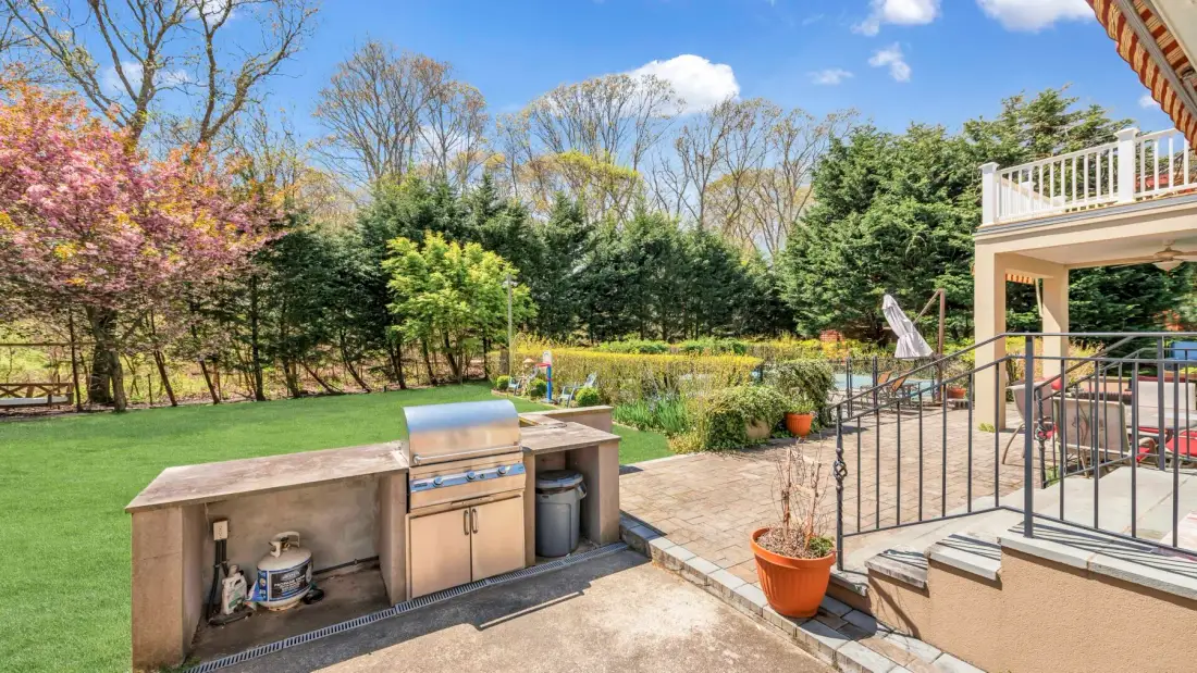 Patio exterior con una parrilla empotrada junto a un césped verde y un jardín cuidado.