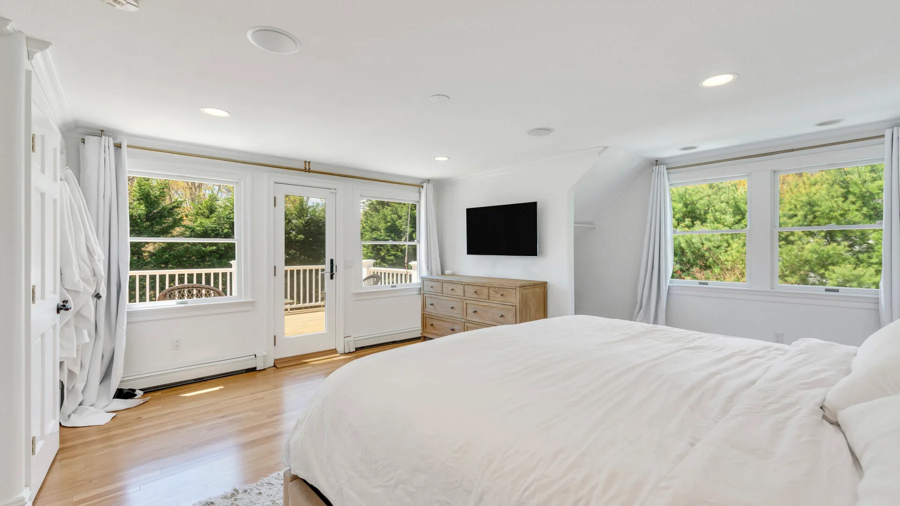 Un dormitorio luminoso con una cama grande, un televisor y ventanas con vistas a la vegetación.