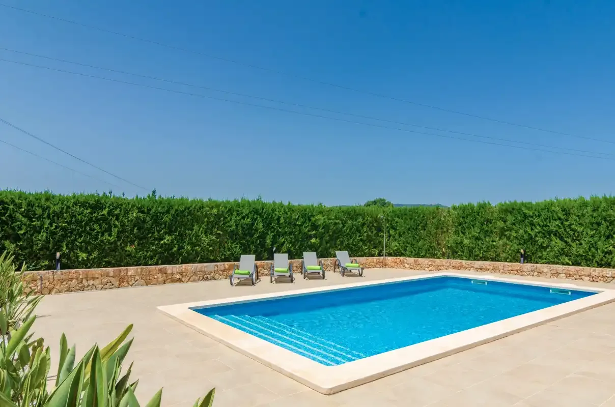 Una piscina al aire libre está junto a un patio pavimentado y un seto alto.
