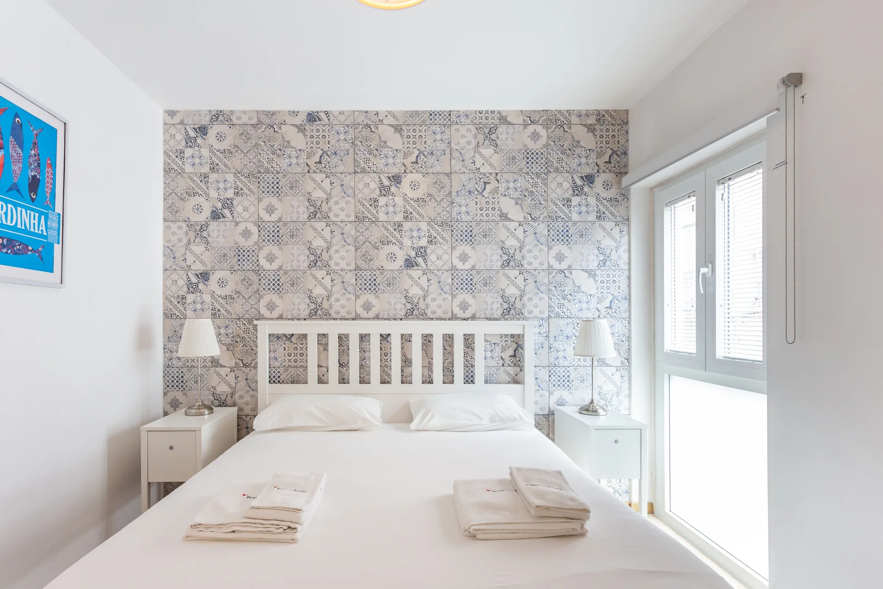 A bright bedroom with a bed against a patterned tile wall and a large window.