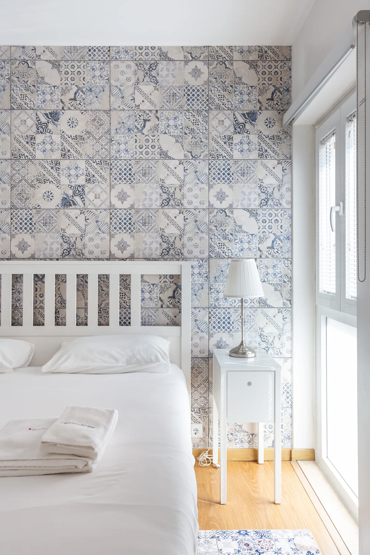 A bedroom with a white bed beside a tall window and patterned wall tiles.