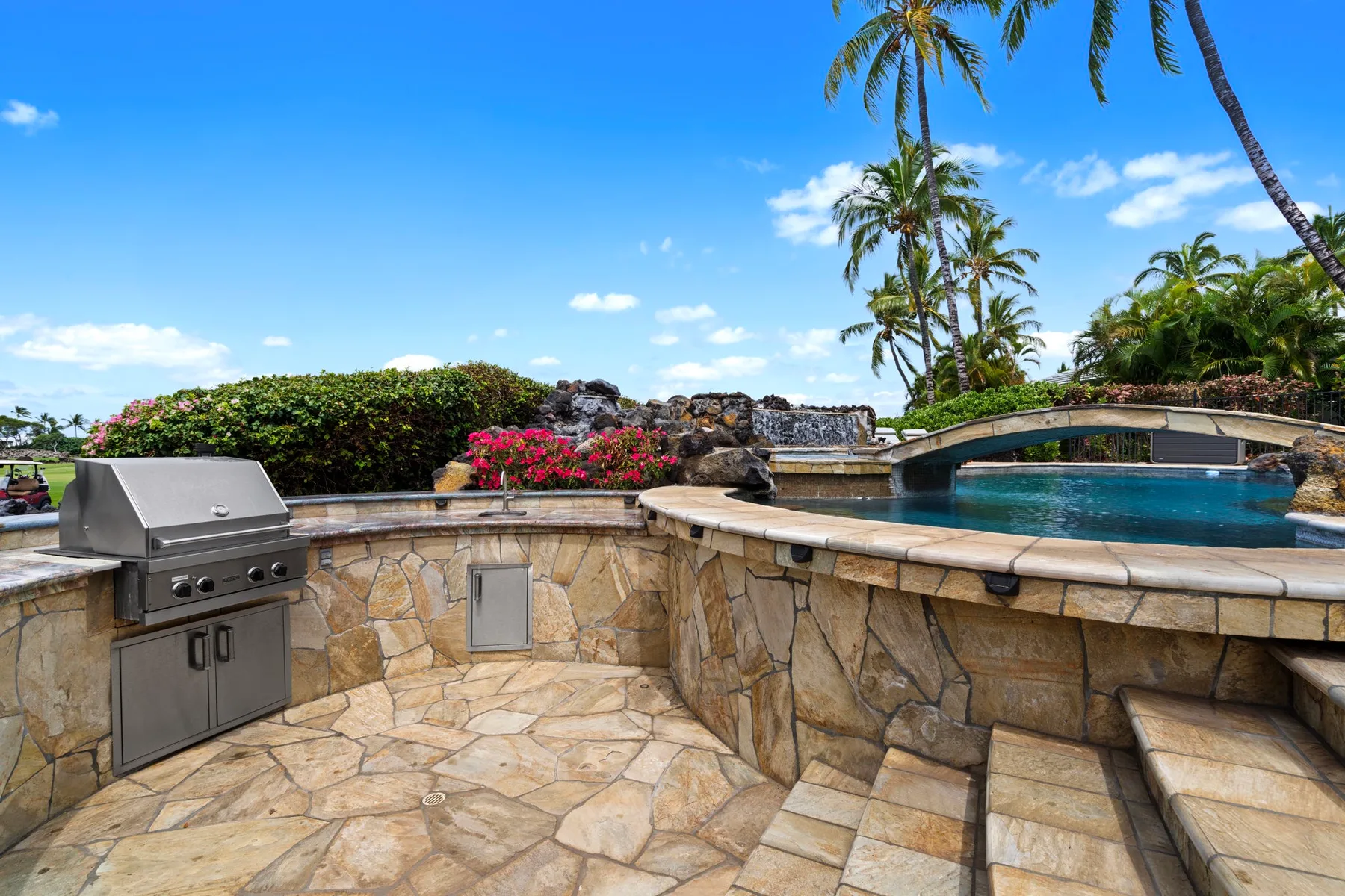 Una piscina tropical al aire libre junto a un patio de piedra y una parrilla empotrada.
