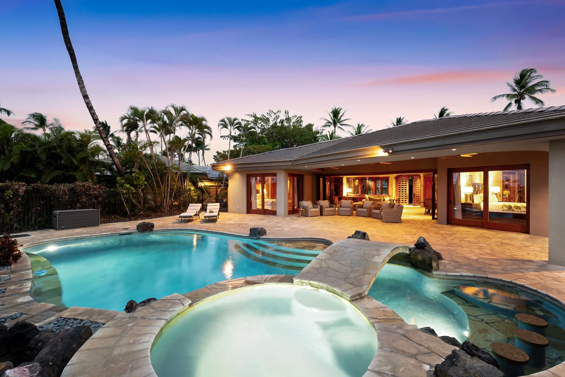 Una casa tropical da a un patio iluminado junto a una piscina y un spa.