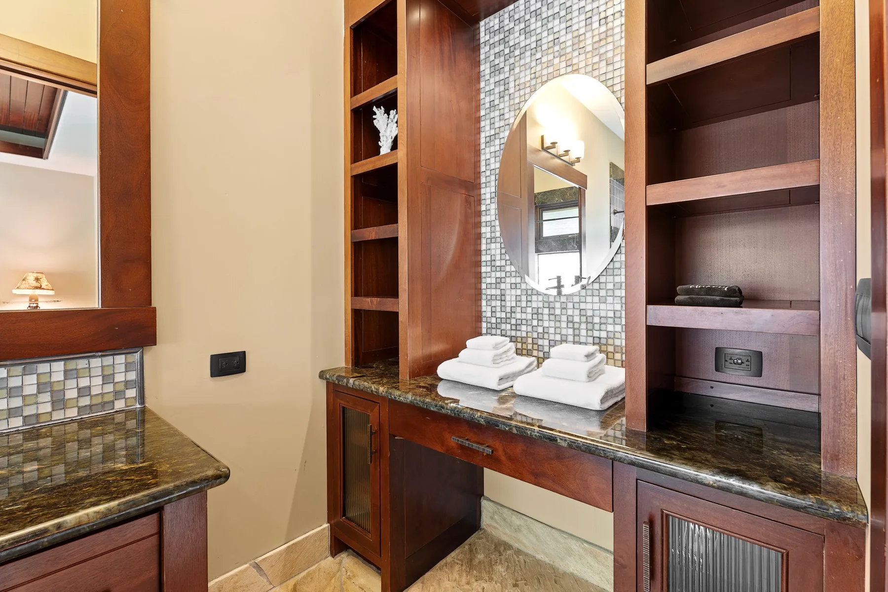 Un tocador de baño con espejo redondo, pared de azulejos y estantes de madera.