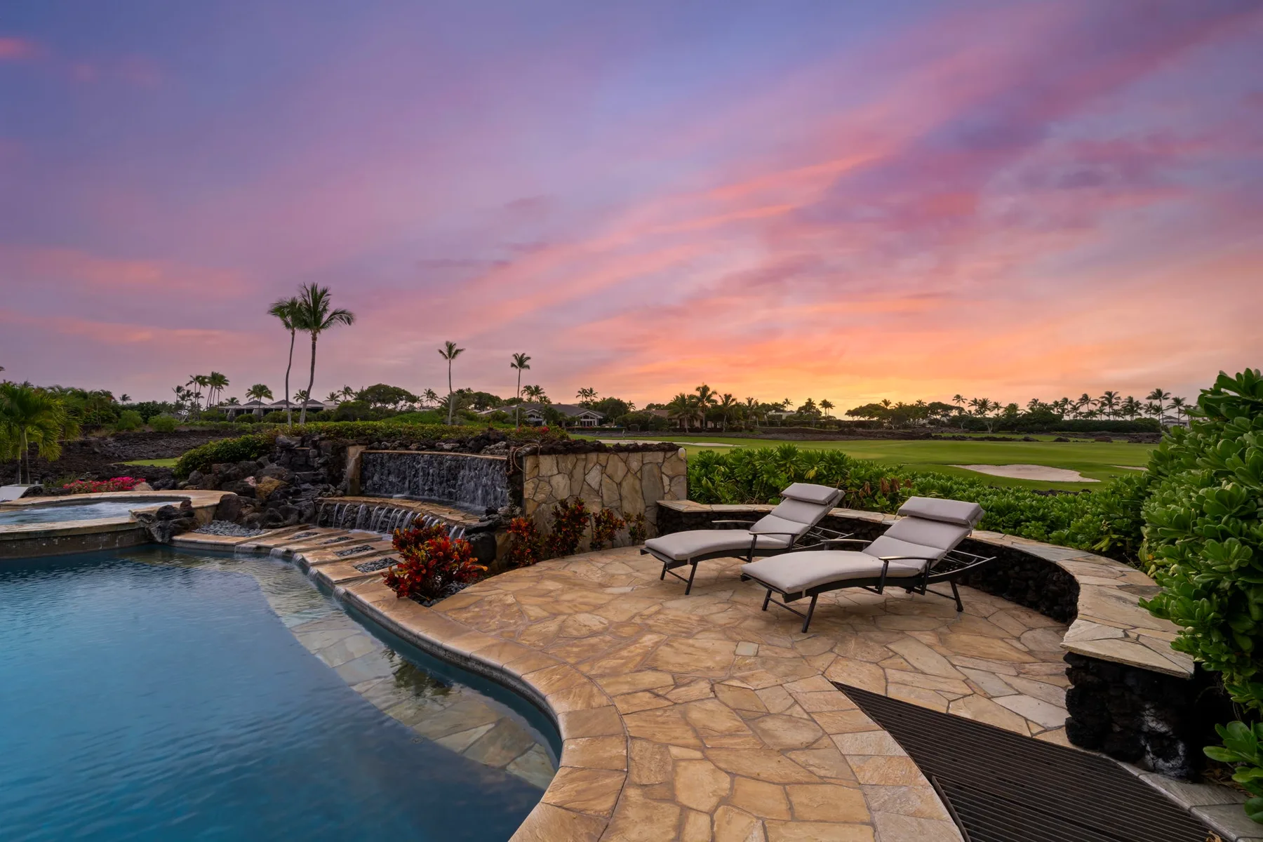 Una piscina exterior y un patio de piedra dan a un paisaje verde bajo un cielo colorido al atardecer.