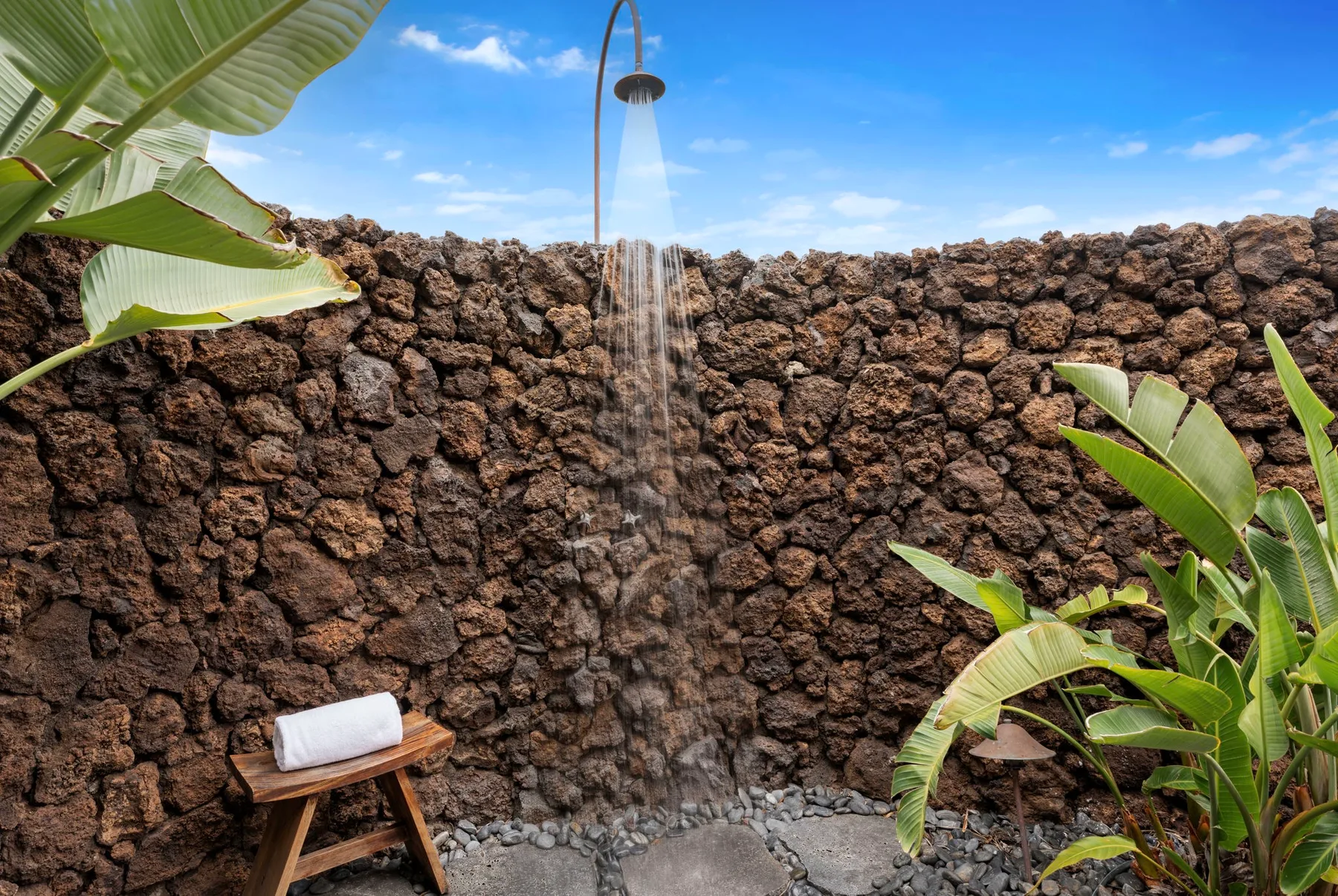 Una ducha al aire libre está frente a un muro de piedra, rodeada de hojas tropicales.