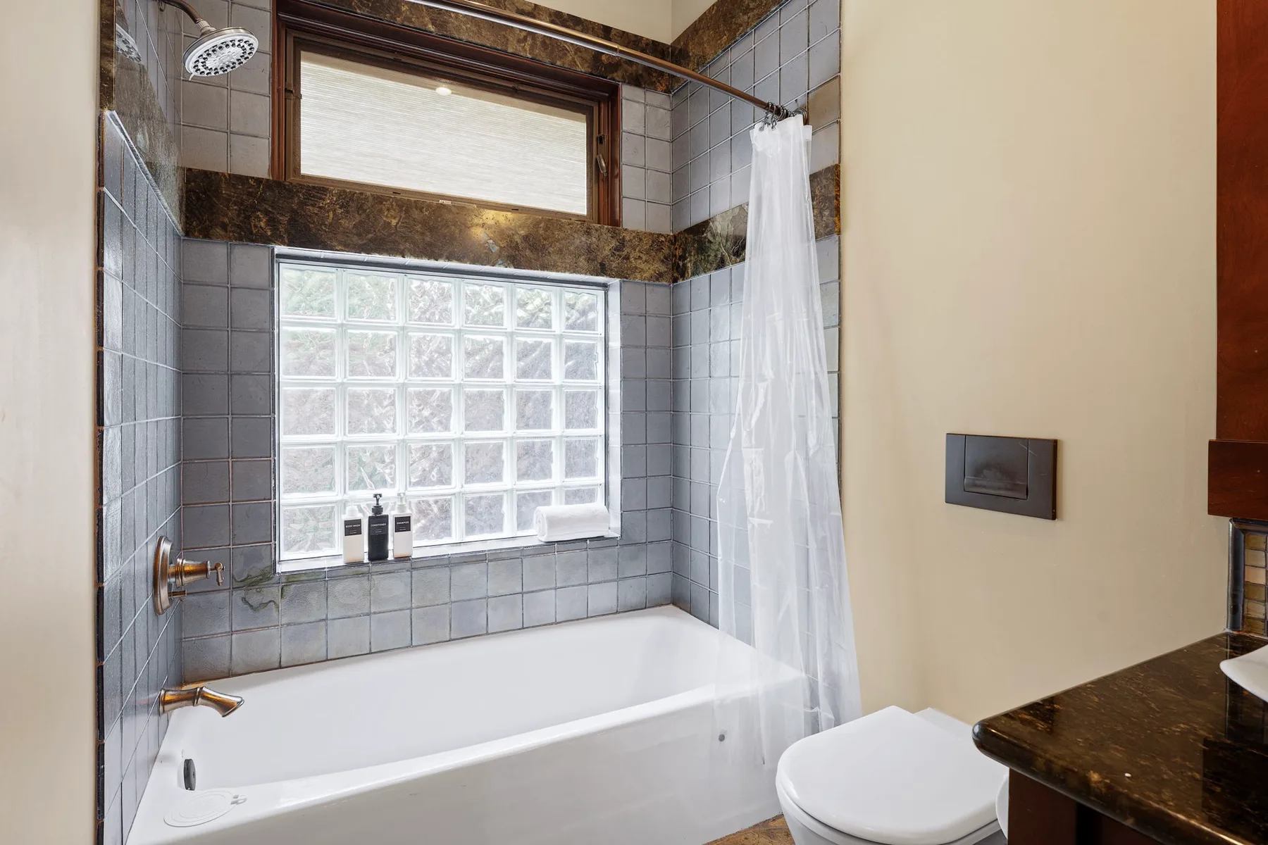 Un baño con una bañera bajo una ventana de bloques de vidrio y una cortina blanca.