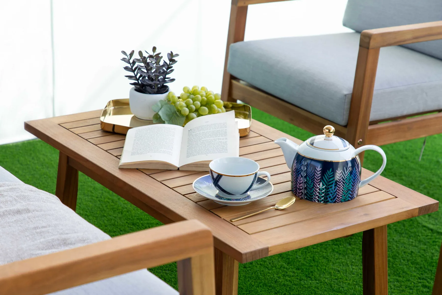Una mesa de patio de madera tiene un libro abierto, una tetera y una taza junto a asientos exteriores.
