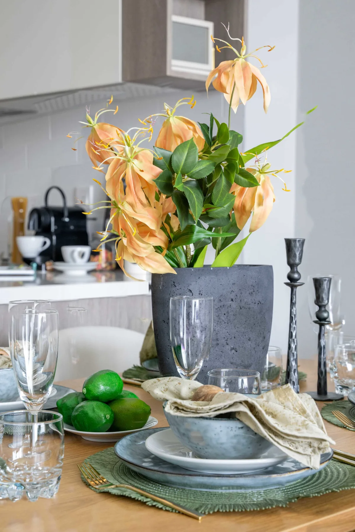 Una mesa de comedor con un arreglo floral y vajilla, con una cocina al fondo.