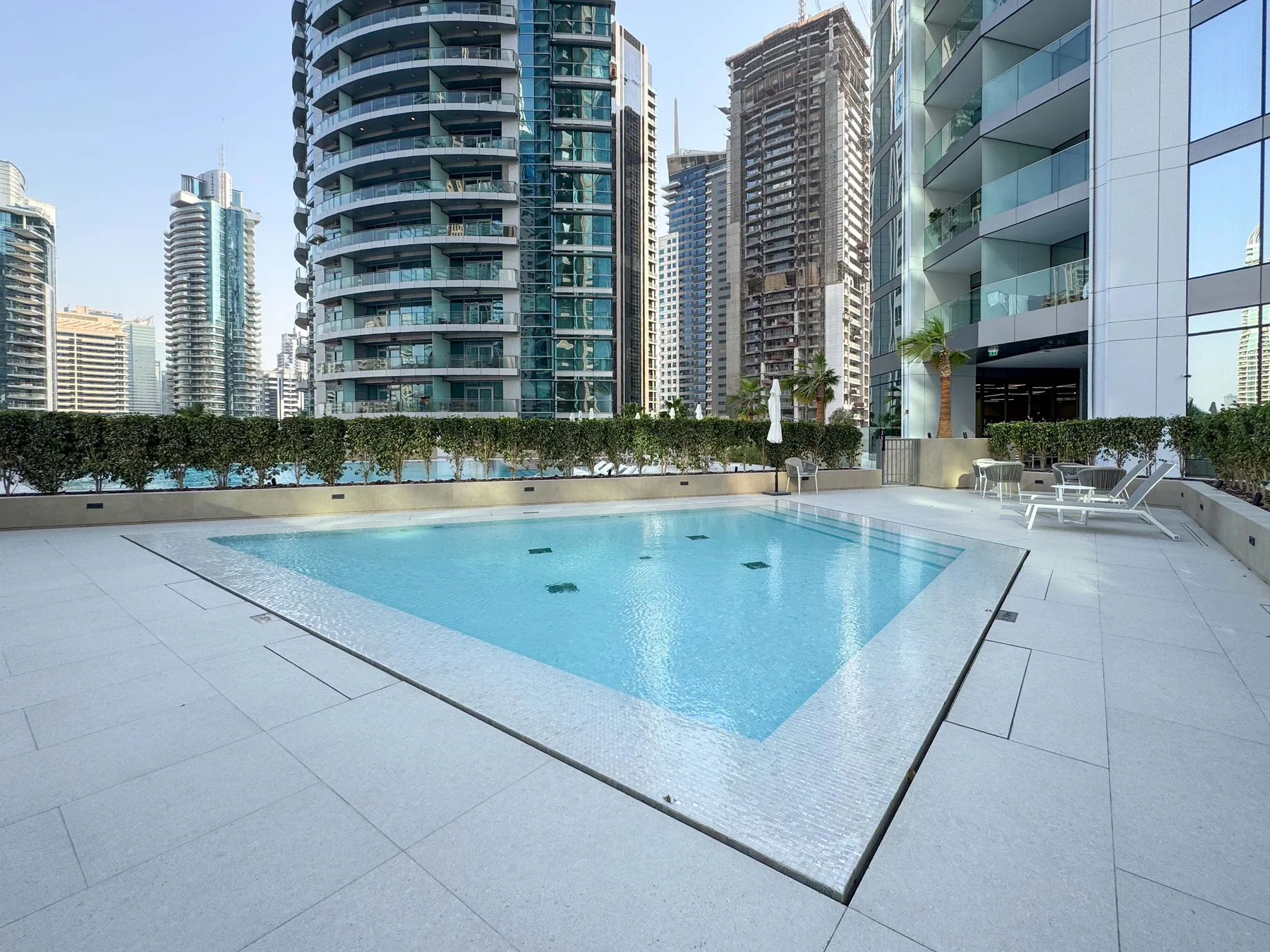 Una piscina al aire libre está rodeada de edificios modernos altos, con tumbonas cerca.