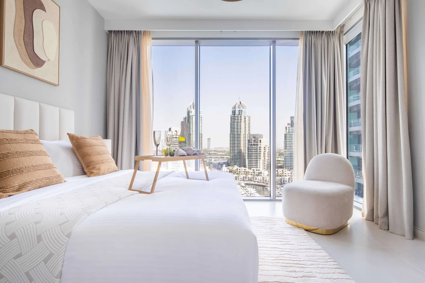 Un dormitorio luminoso con una cama, un sillón redondeado y vistas a la ciudad por grandes ventanales.
