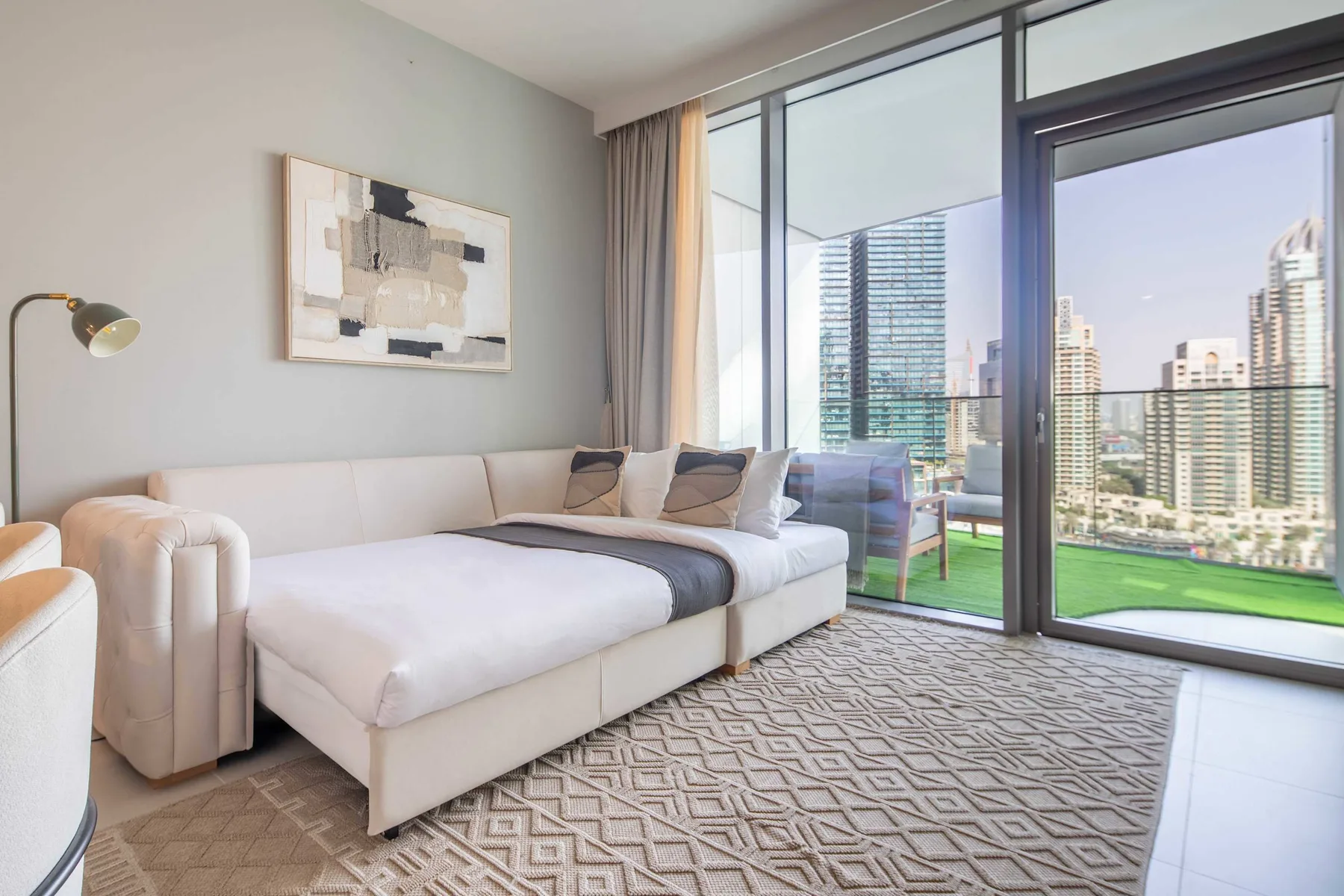 Un dormitorio luminoso con una cama grande junto a ventanales con vistas a edificios urbanos.
