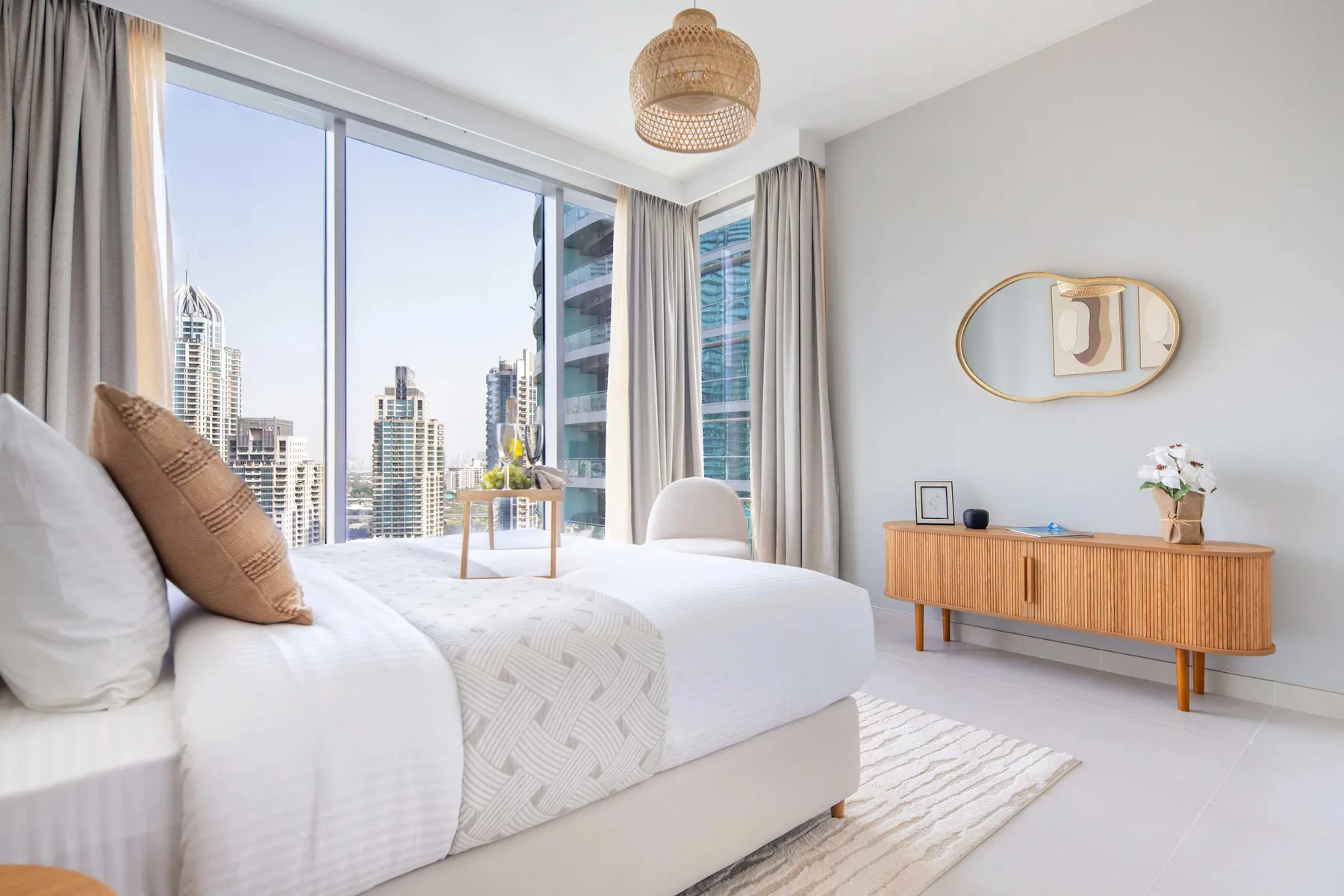 Un dormitorio luminoso con una cama grande junto a ventanales con vistas a edificios urbanos.