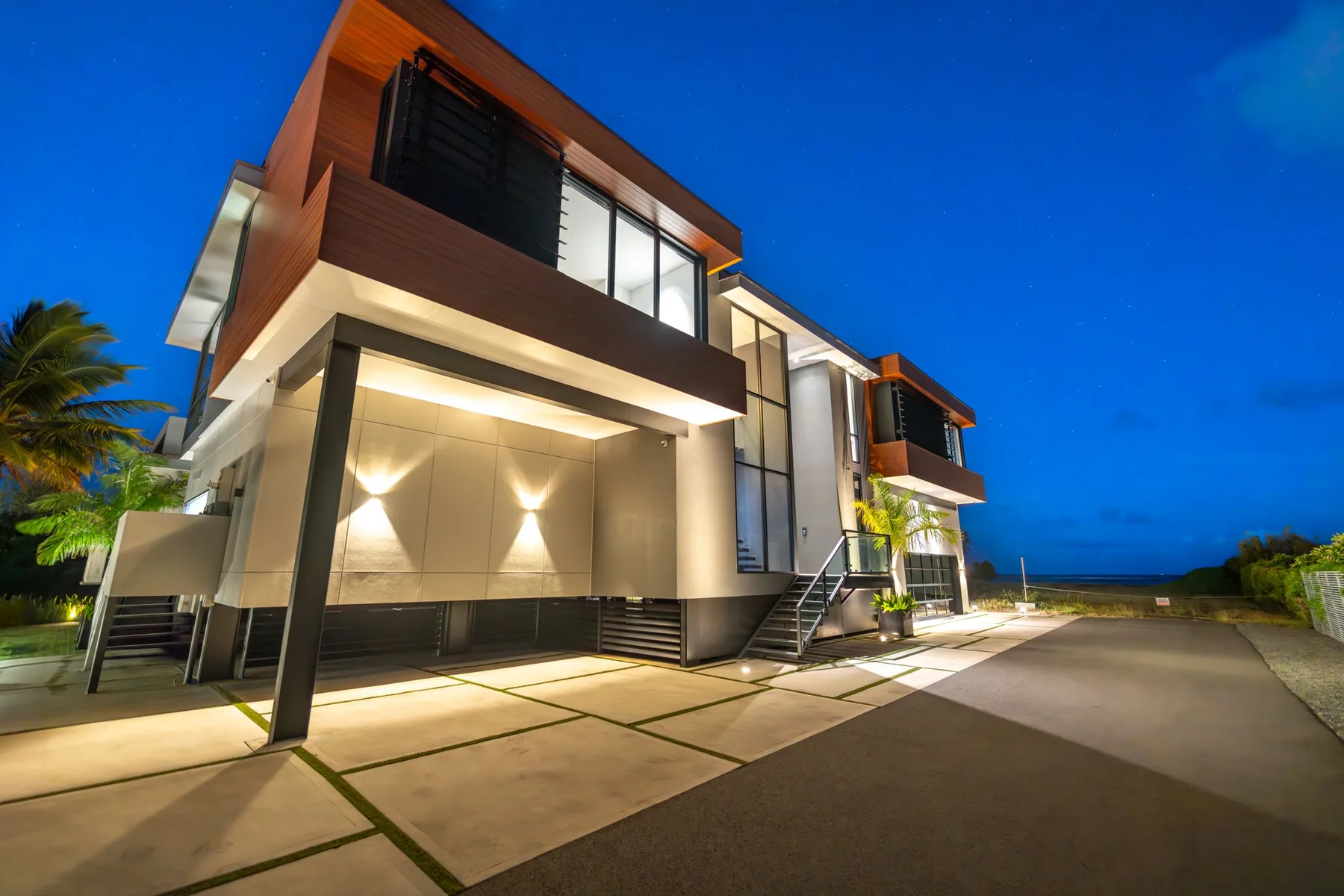 Exterior de una casa moderna al anochecer, con ventanas iluminadas y una entrada pavimentada.