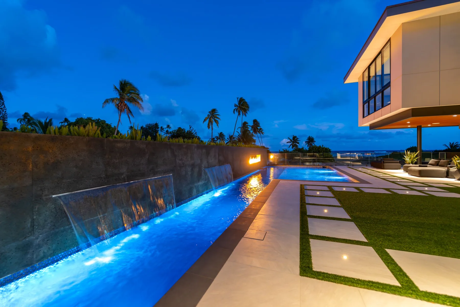 Una piscina exterior iluminada junto a un patio pavimentado al anochecer.