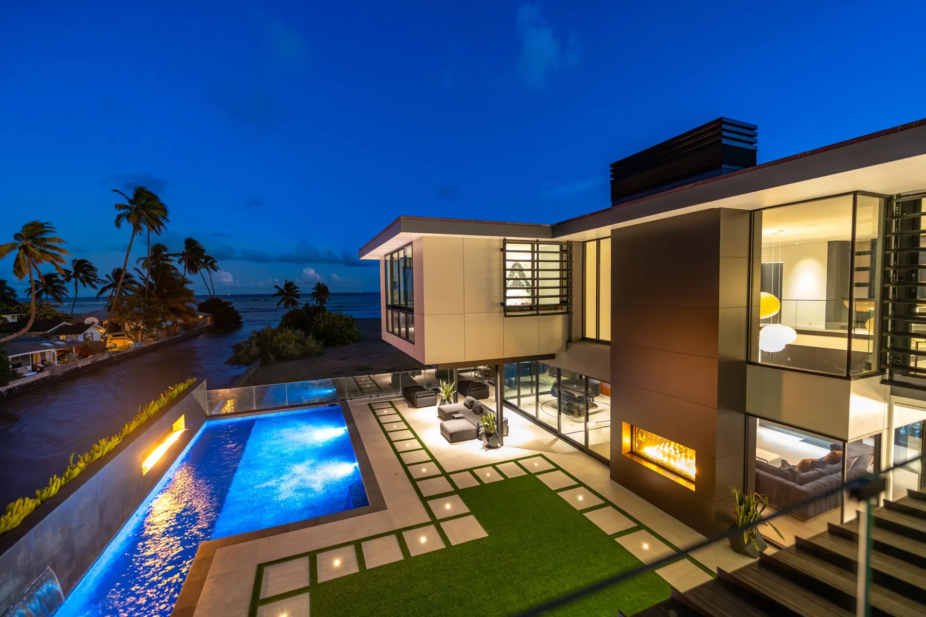 Una casa moderna da a una piscina iluminada y al océano al anochecer.