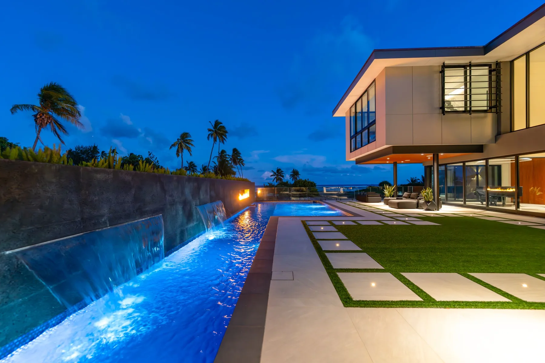 Una casa moderna junto a una piscina iluminada y un césped ajardinado al anochecer.